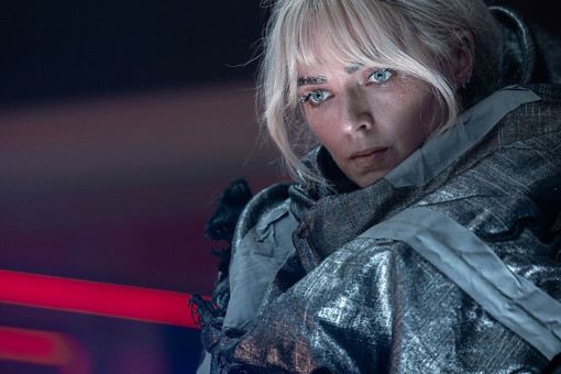 A woman with blonde hair and blue eyes, wrapped in mylar blankets duck taped around her cold body covered in frost, stares intently at something off screen