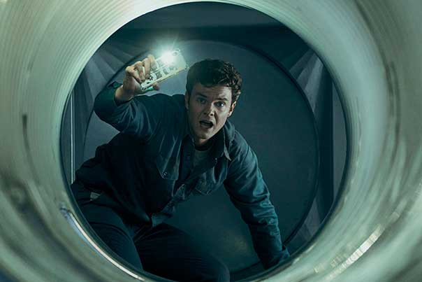 The Boys star Jack Quaid crouched in a metallic tunnel, holding his phone and shining its flashlight.