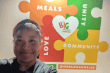 A volunteer taking a selfie in front of a wall that reads "meals, love, community, unity" at Our Big Kitchen